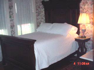 Photo of a queen bed in one of the inn's rooms, with an antique lamp, nightstand and wallpaper.